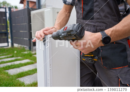 Man fastens white drawer side with cordless drill near grass outside Man fastens white drawer side with cordless drill near grass outside 139811270