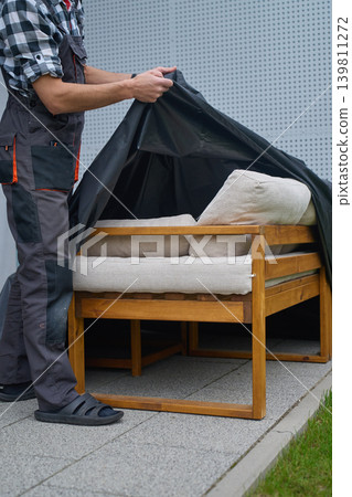 Man removes protective cover from outdoor sofa with cushions on patio. Concept of garden furniture care Man removes protective cover from outdoor sofa with cushions on patio. Concept of garden furniture care 139811272