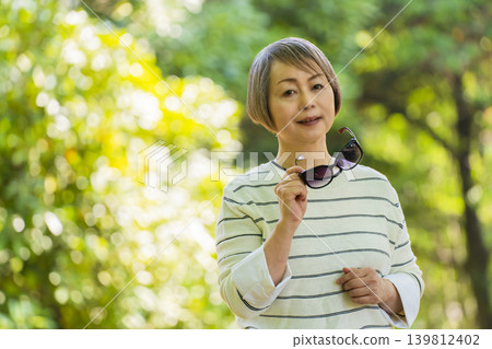 一位戴著太陽眼鏡的老婦人在綠樹環繞的公園散步。 一位戴著太陽眼鏡的老婦人在綠樹環繞的公園散步。 139812402