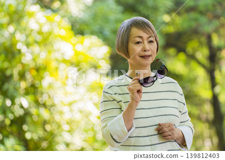 A senior woman wearing sunglasses takes a stroll in a park surrounded by fresh greenery. 139812403