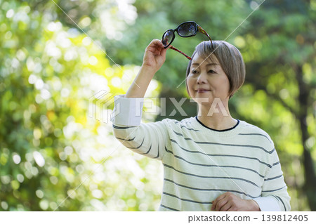 一位戴著太陽眼鏡的老婦人在綠樹環繞的公園散步。 一位戴著太陽眼鏡的老婦人在綠樹環繞的公園散步。 139812405