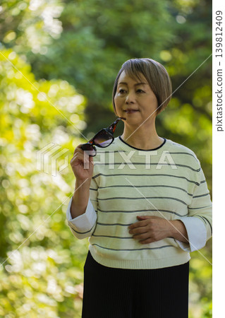 一位戴著太陽眼鏡的老婦人在綠樹環繞的公園散步。 一位戴著太陽眼鏡的老婦人在綠樹環繞的公園散步。 139812409