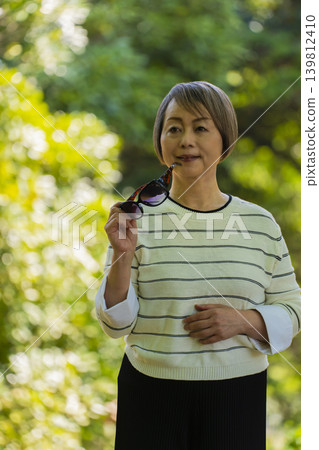 A senior woman wearing sunglasses takes a stroll in a park surrounded by fresh greenery. 139812410