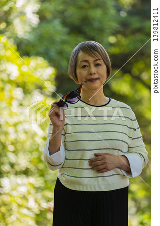 一位戴著太陽眼鏡的老婦人在綠樹環繞的公園散步。 一位戴著太陽眼鏡的老婦人在綠樹環繞的公園散步。 139812411