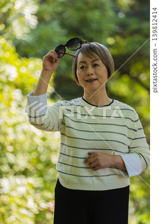 A senior woman wearing sunglasses takes a stroll in a park surrounded by fresh greenery. A senior woman wearing sunglasses takes a stroll in a park surrounded by fresh greenery. 139812414