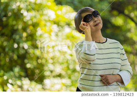 A senior woman wearing sunglasses takes a stroll in a park surrounded by fresh greenery. A senior woman wearing sunglasses takes a stroll in a park surrounded by fresh greenery. 139812423