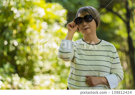 一位戴著太陽眼鏡的老婦人在綠樹環繞的公園散步。 一位戴著太陽眼鏡的老婦人在綠樹環繞的公園散步。 139812424