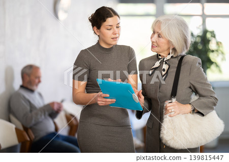 Positive polite secretary or social worker talking to aged woman visitor in reception area in background queue of people 139815447