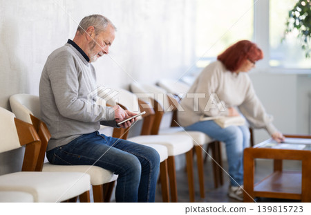 Elderly man sitting in clinic waiting area scrolling mobile phone 139815723