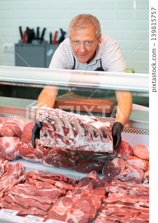Positive elderly salesman in apron and disposable gloves shows raw beef tomahawk steak Positive elderly salesman in apron and disposable gloves shows raw beef tomahawk steak 139815757