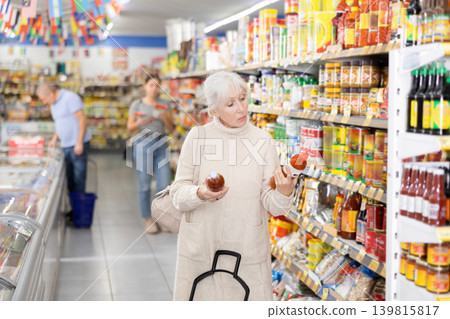 Mature woman choosing hot chili sauce for herself in supermarket Mature woman choosing hot chili sauce for herself in supermarket 139815817