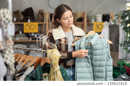 Young girl choosing light green vest for autumn-winter in clothing store Young girl choosing light green vest for autumn-winter in clothing store 139815818