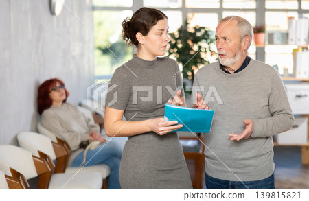Positive polite nurse or doctor talking to aged man patient in reception area in background queue of people Positive polite nurse or doctor talking to aged man patient in reception area in background queue of people 139815821