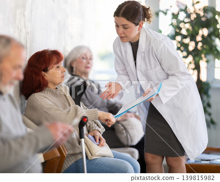Polite young nurse or doctor talking to mature woman patient standing in waiting room of medical clinic, with her back to visitors sitting in chairs Polite young nurse or doctor talking to mature woman patient standing in waiting room of medical clinic, with her back to visitors sitting in chairs 139815982
