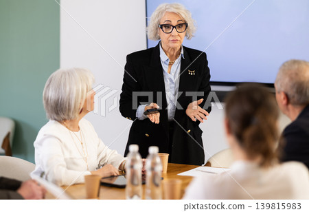Mature female manager with remote control addresses round table meeting participants Mature female manager with remote control addresses round table meeting participants 139815983