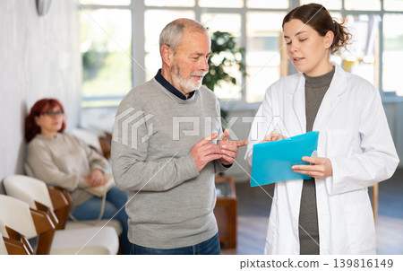 Polite young nurse or doctor talking to mature man patient standing in waiting room of medical clinic, with her back to visitors sitting in chairs Polite young nurse or doctor talking to mature man patient standing in waiting room of medical clinic, with her back to visitors sitting in chairs 139816149