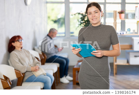 Portrait of young female secretary standing with folder with documents and materials in hands and inviting in cabinet or to appointment Portrait of young female secretary standing with folder with documents and materials in hands and inviting in cabinet or to appointment 139816174