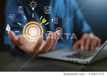 Business goal target success with dartboard and trend graph marketing and research icons over hand while person uses laptop for corporate strategy and business target planning Business goal target success with dartboard and trend graph marketing and research icons over hand while person uses laptop for corporate strategy and business target planning 139817375