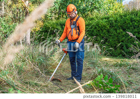 Man using a string trimmer to cut weeds and tall grass in an overgrown field during autumn in a Man using a string trimmer to cut weeds and tall grass in an overgrown field during autumn in a 139818011