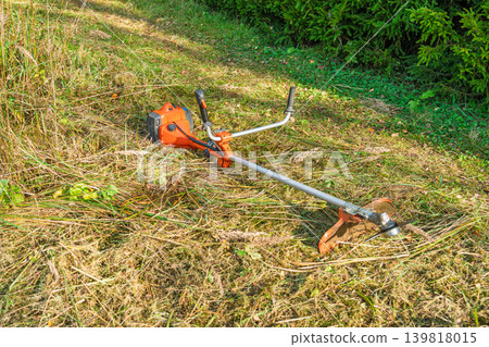 Orange string trimmer resting on freshly mown grass, garden care equipment set out on the lawn Orange string trimmer resting on freshly mown grass, garden care equipment set out on the lawn 139818015