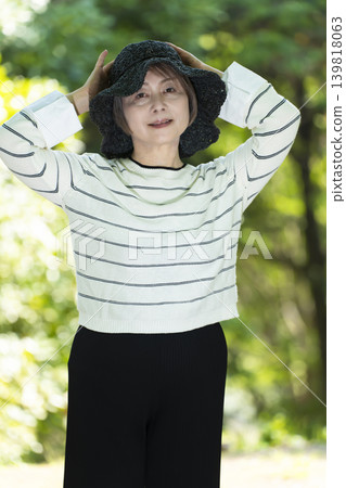 A senior woman wearing a hat takes a stroll in a park surrounded by fresh greenery. A senior woman wearing a hat takes a stroll in a park surrounded by fresh greenery. 139818063