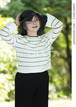 A senior woman wearing a hat takes a stroll in a park surrounded by fresh greenery. A senior woman wearing a hat takes a stroll in a park surrounded by fresh greenery. 139818064