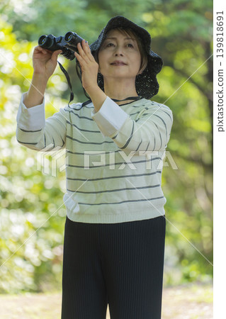 一位老婦人在綠樹環繞的公園裡用望遠鏡觀鳥。 139818691