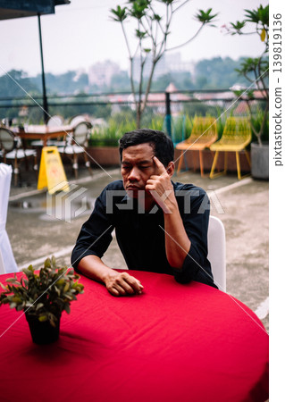 Southeast Asian man thinking while holding head at outdoor round table Southeast Asian man thinking while holding head at outdoor round table 139819136