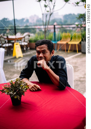Southeast Asian man contemplating with hand on mouth at outdoor table 139819140