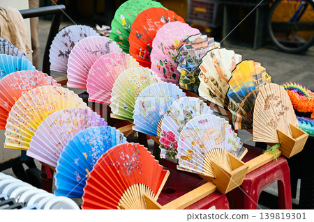 Traditional colorful handmade folding bamboo fans with painted patterns displayed at outdoor souvenir street market booth in Vietnam Traditional colorful handmade folding bamboo fans with painted patterns displayed at outdoor souvenir street market booth in Vietnam 139819301