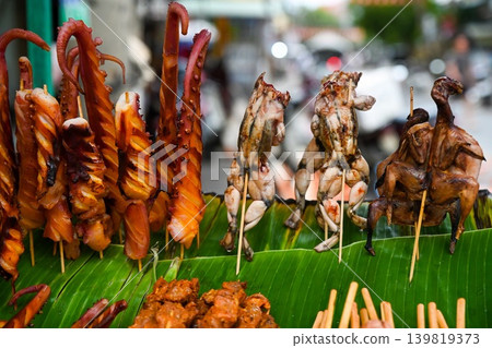 Exotic grilled street food including giant octopus tentacles frog skewers and whole birds displayed on green banana leaf at outdoor market in Vietnam Exotic grilled street food including giant octopus tentacles frog skewers and whole birds displayed on green banana leaf at outdoor market in Vietnam 139819373