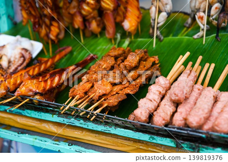Assorted traditional grilled meat skewers and pork meatballs nem nuong displayed on green banana leaf at outdoor street food market in Vietnam Assorted traditional grilled meat skewers and pork meatballs nem nuong displayed on green banana leaf at outdoor street food market in Vietnam 139819376
