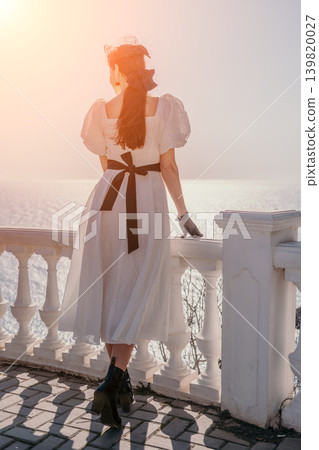 Woman white dress ocean standing on a balcony overlooks the shimmering sea during a peaceful golden hour sunset Woman white dress ocean standing on a balcony overlooks the shimmering sea during a peaceful golden hour sunset 139820027