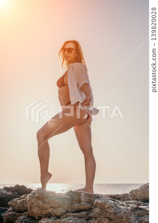Woman beach sunset standing on coastal rocks wearing a white shirt and bikini during golden hour with copy space Woman beach sunset standing on coastal rocks wearing a white shirt and bikini during golden hour with copy space 139820028