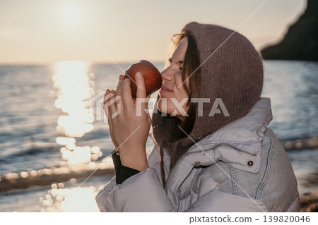 Woman apple beach smelling fresh fruit by the sea during a golden sunset while wearing warm winter clothing and a hood 139820046
