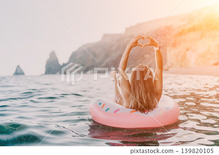 Woman, donut, ocean. Relaxed woman on inflatable donut making heart gesture in sea with copy space. Woman, donut, ocean. Relaxed woman on inflatable donut making heart gesture in sea with copy space. 139820105