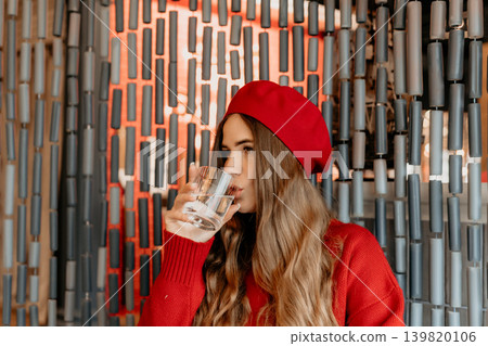Woman drinking water wearing a red beret and sweater inside a cafe against a unique artistic cylinder wall backdrop 139820106