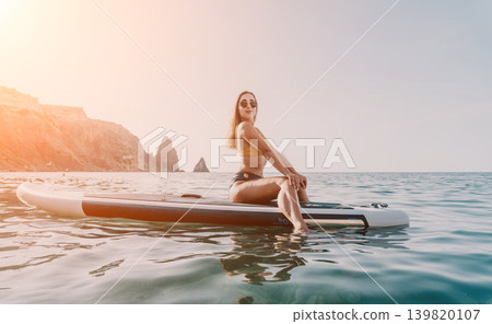 Woman Paddleboard Ocean - Young woman in a bikini sits on a paddleboard in the ocean on a sunny day. 139820107