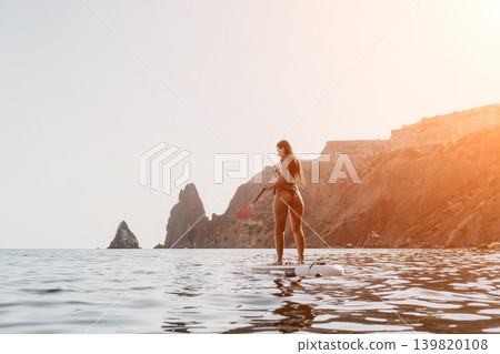 Woman Paddleboarding Ocean Coastline - A woman enjoys a day of paddleboarding in the ocean, with a mountainous coastline in the background. 139820108