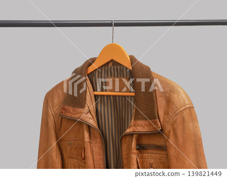 Brown leather jacket hanging on hanger below rail. Outerwear garment displayed on clothing rack. Menswear wardrobe option for transitional weather outfits.. Brown leather jacket hanging on hanger below rail. Outerwear garment displayed on clothing rack. Menswear wardrobe option for transitional weather outfits.. 139821449
