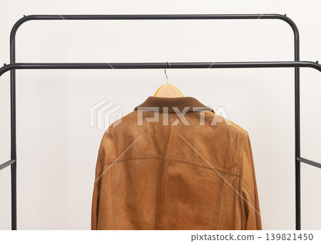 Brown leather jacket hanging on metal rack. Outerwear garment placed on wooden hanger. Menswear wardrobe choice for autumn spring season.. Brown leather jacket hanging on metal rack. Outerwear garment placed on wooden hanger. Menswear wardrobe choice for autumn spring season.. 139821450