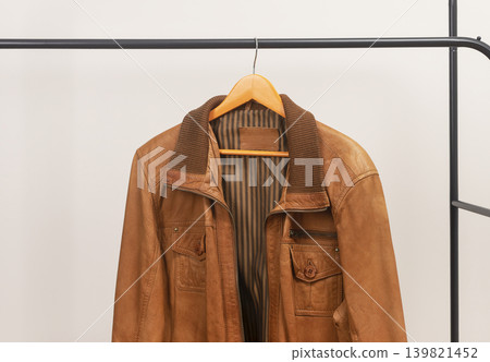 Brown leather jacket hanging on rack with zipper. Outerwear garment placed on wooden hanger. Menswear wardrobe clothing for autumn spring season.. Brown leather jacket hanging on rack with zipper. Outerwear garment placed on wooden hanger. Menswear wardrobe clothing for autumn spring season.. 139821452