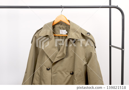 Khaki women trench coat hanging on metal rack. Outerwear garment placed on wooden hanger. Female wardrobe piece with military style inspiration. Khaki women trench coat hanging on metal rack. Outerwear garment placed on wooden hanger. Female wardrobe piece with military style inspiration. 139821518