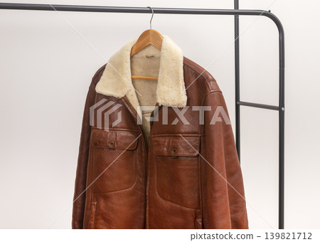 Brown leather jacket hangs on wooden hanger against white background. Shearling collar outerwear rests on black metal rack creating wardrobe display. Minimalist retail fashion concept for Brown leather jacket hangs on wooden hanger against white background. Shearling collar outerwear rests on black metal rack creating wardrobe display. Minimalist retail fashion concept for 139821712