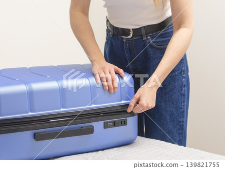 Woman closing suitcase on bed during travel preparation. Indoor packing moment with clothes and hand Woman closing suitcase on bed during travel preparation. Indoor packing moment with clothes and hand 139821755