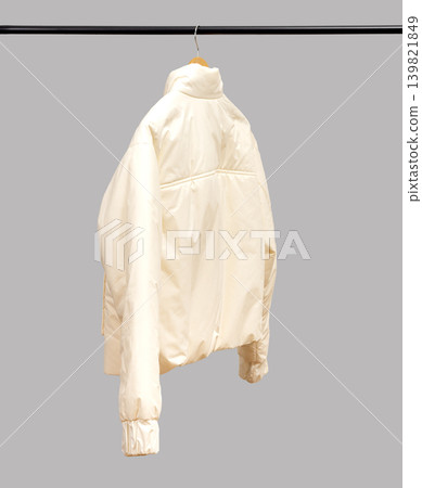Beige jacket hanging on black metal rack against gray wall. Back view garment suspended on hanger. Fashion storage concept for retail, wardrobe, clothing.. Beige jacket hanging on black metal rack against gray wall. Back view garment suspended on hanger. Fashion storage concept for retail, wardrobe, clothing.. 139821849
