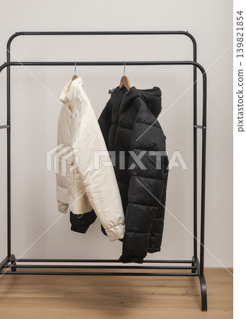 Beige and black puffer jackets hang on metal rack against white background. Quilted outerwear rests on wooden hangers forming retail wardrobe display. Minimalist fashion concept for boutique Beige and black puffer jackets hang on metal rack against white background. Quilted outerwear rests on wooden hangers forming retail wardrobe display. Minimalist fashion concept for boutique 139821854