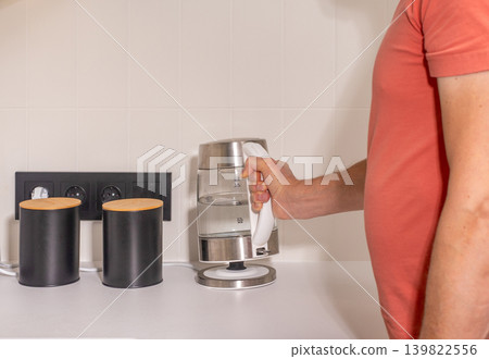Adult man places electric kettle near kitchen containers on counter. Mature male prepares hot water appliance during everyday home routine. Simple kitchen preparation activity in modern 139822556