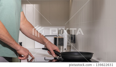 Mature man holds frying pan while preparing food at kitchen stove. Adult male cooks meal during everyday home routine. Simple domestic cooking moment inside modern kitchen interior.. 139822631