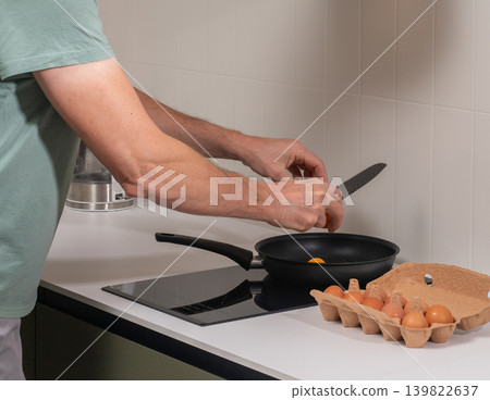 Mature man cracks egg into frying pan while cooking at kitchen stove. Adult male prepares breakfast meal during everyday domestic routine. Home food preparation activity in modern kitchen Mature man cracks egg into frying pan while cooking at kitchen stove. Adult male prepares breakfast meal during everyday domestic routine. Home food preparation activity in modern kitchen 139822637